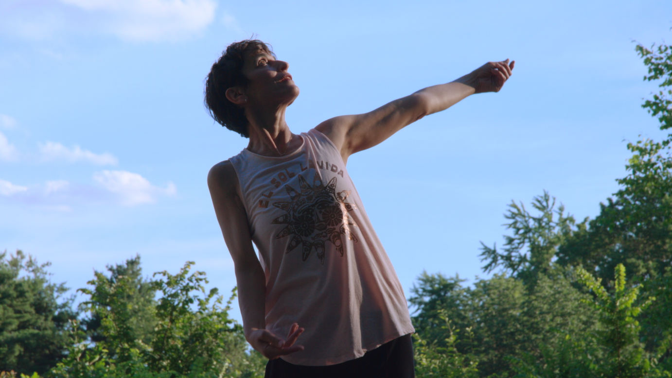 A wiry woman sporting short dark hair, sways to her right with her left arm lifted horizontally. She wears a light pink, no sleeved shirt that hits her thighs. She is wearing black pants. Her look is serene. She dances in front of a light blue sky. .